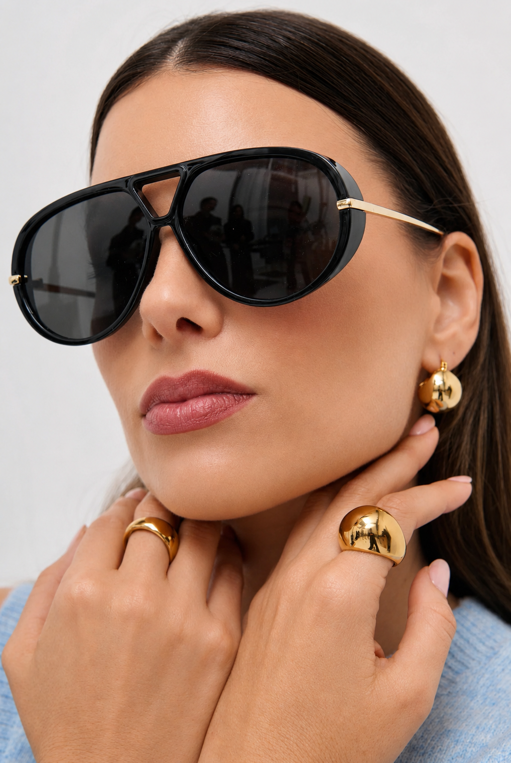 A close-up of a woman wearing oversized black aviator-style sunglasses with gold metal temples, along with gold hoop earrings and gold rings, against a light gray background.