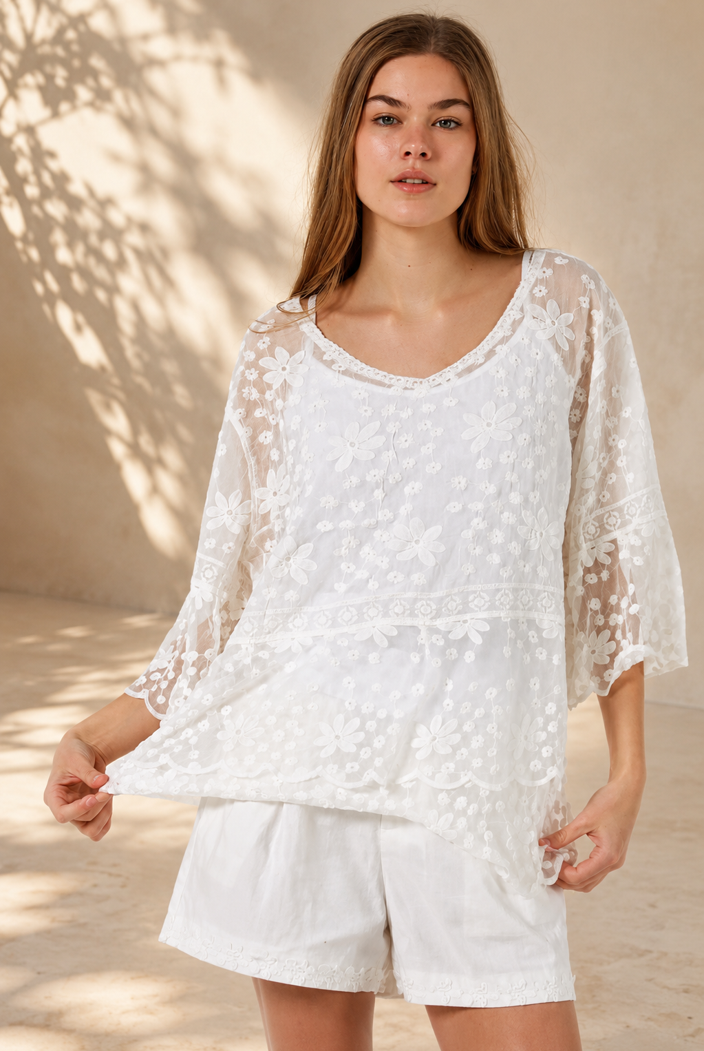 A woman wearing a white lace long-sleeve tunic top with matching white shorts, standing in a sunlit outdoor setting with a beige wall and potted plant in the background.