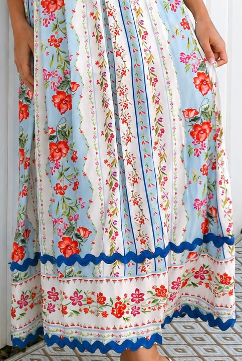 A woman wearing a sleeveless, A-line maxi dress with a white base, blue scalloped trim, and a red and pink floral print, standing outdoors in front of green plants.
