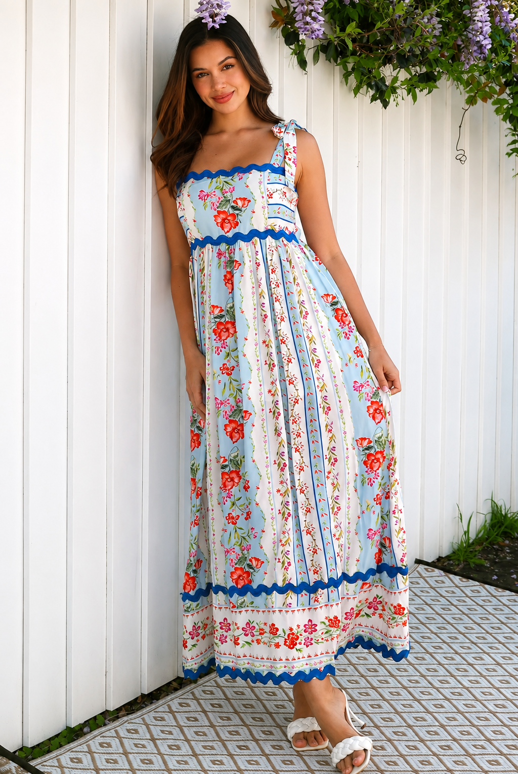 A woman wearing a sleeveless, A-line maxi dress with a white base, blue scalloped trim, and a red and pink floral print, standing outdoors in front of green plants.