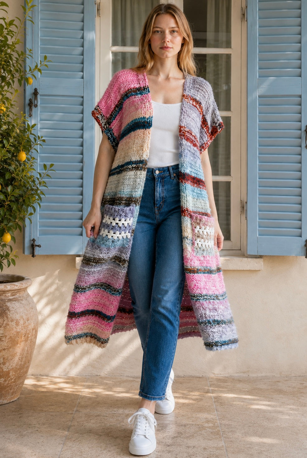 A woman wearing a long, open-front, short-sleeved chunky knit cardigan with horizontal multicolor stripes in pink, blue, beige, brown, and white, paired with a white tank top and blue jeans, standing in front of a window with blue shutters.