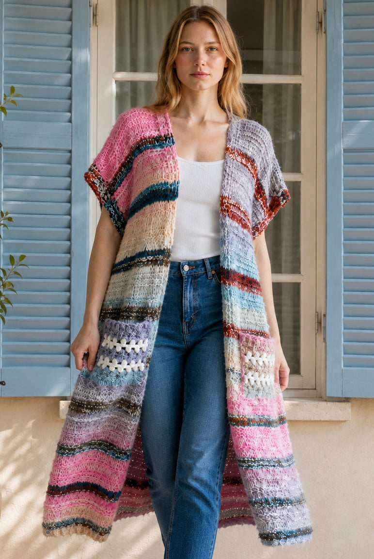 A woman wearing a long, open-front, short-sleeved chunky knit cardigan with horizontal multicolor stripes in pink, blue, beige, brown, and white, paired with a white tank top and blue jeans, standing in front of a window with blue shutters.