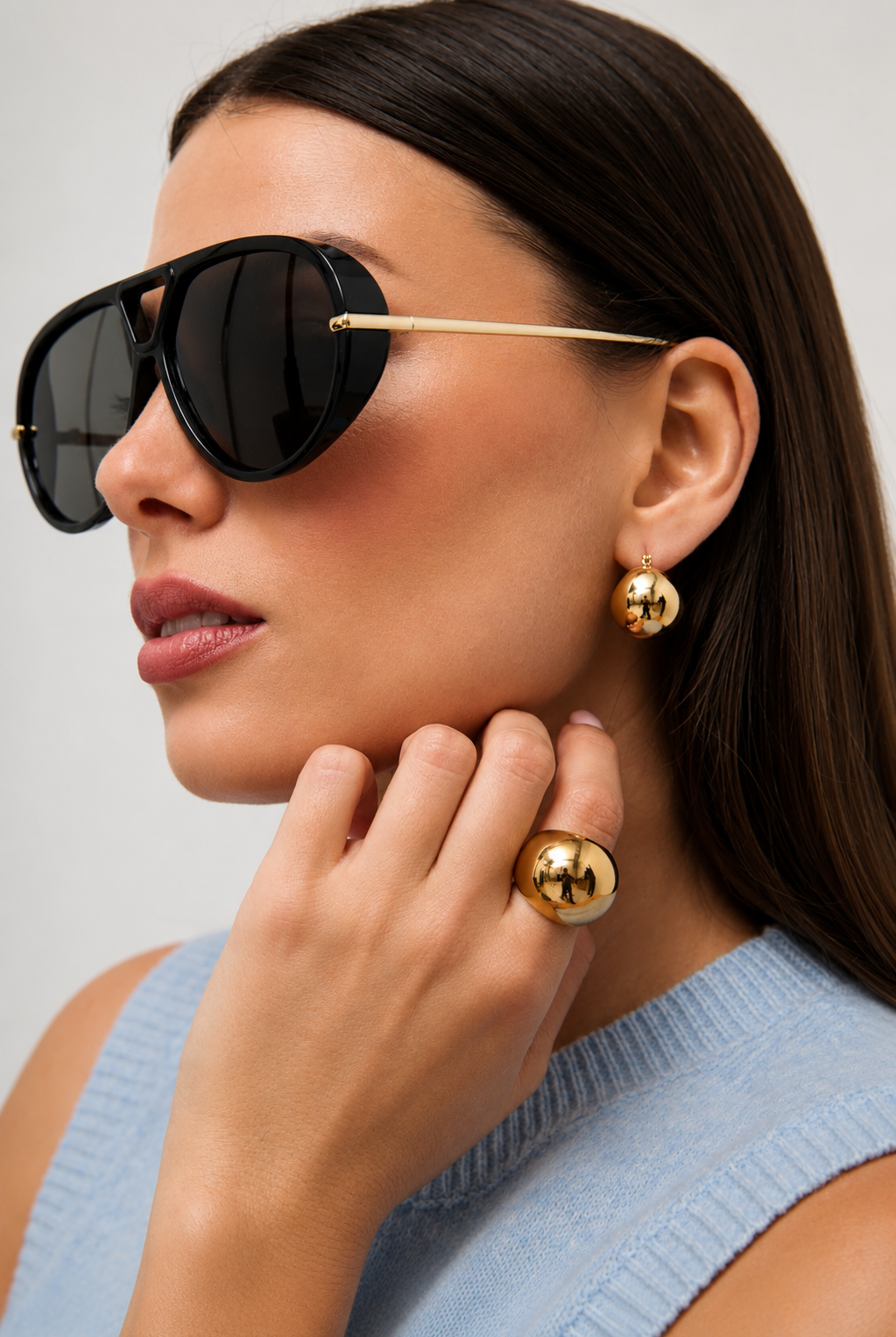 A close-up of a woman wearing oversized black aviator-style sunglasses with gold metal temples, along with gold hoop earrings and gold rings, against a light gray background.