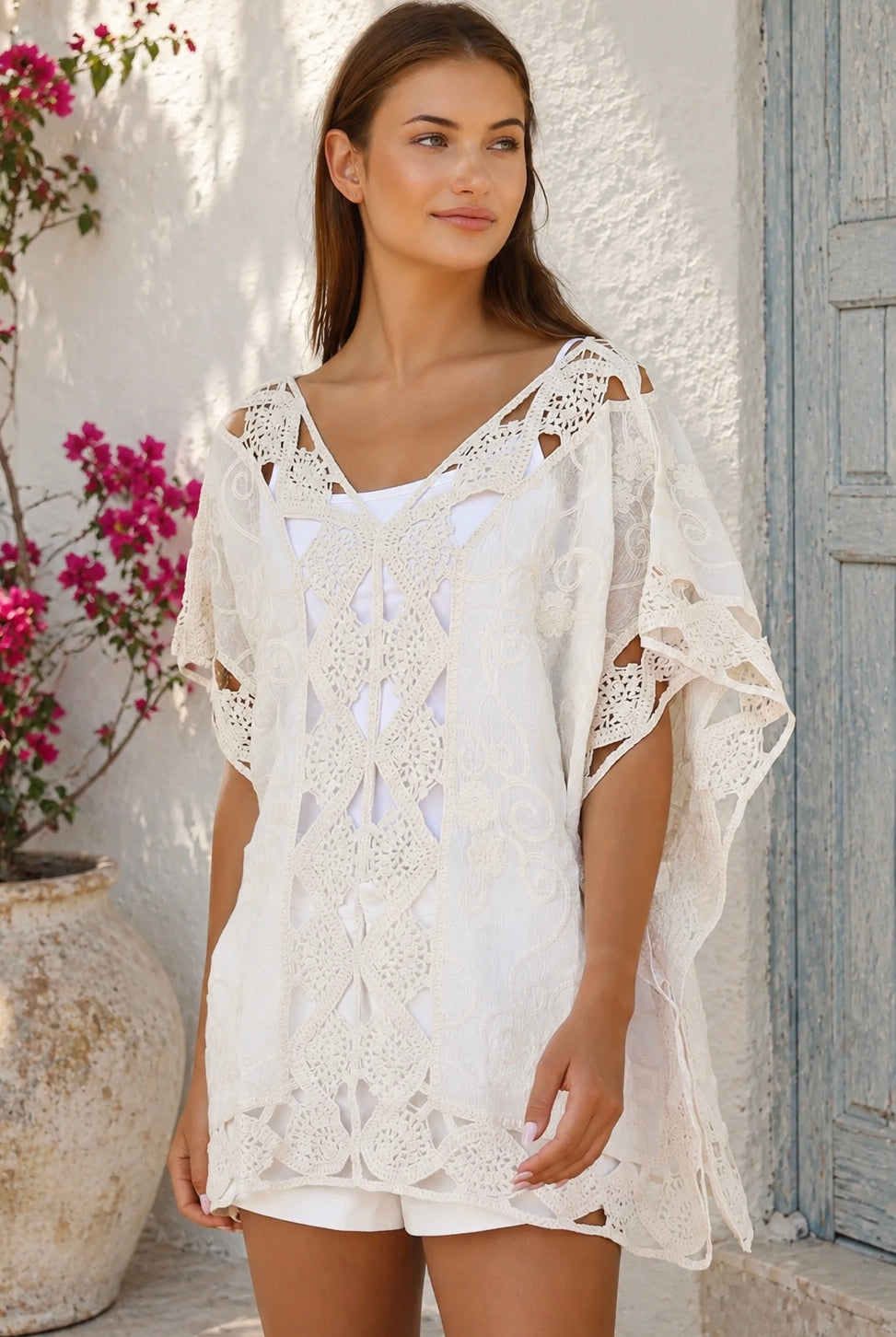 Cream crochet boho kaftan with ornate lace panels and relaxed sleeves in a sunlit Mediterranean coastal setting.