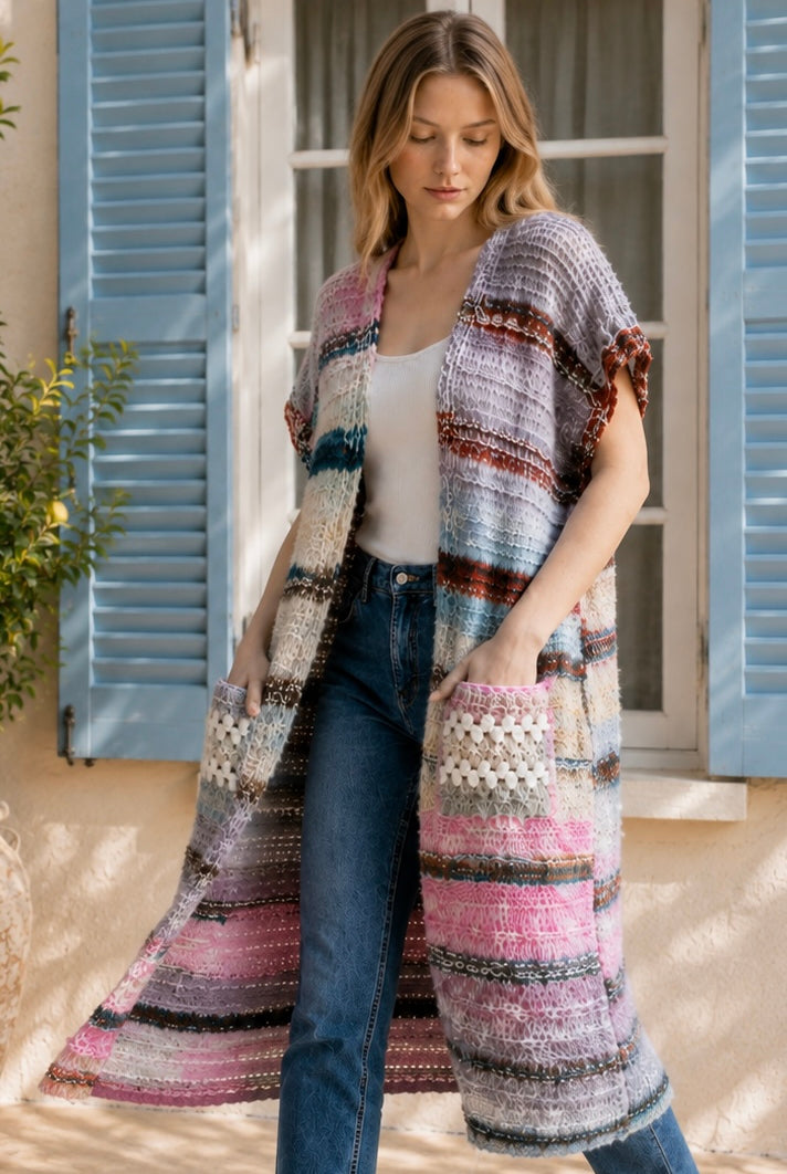 A woman wearing a long, open-front, short-sleeved chunky knit cardigan with horizontal multicolor stripes in pink, blue, beige, brown, and white, paired with a white tank top and blue jeans, standing in front of a window with blue shutters.