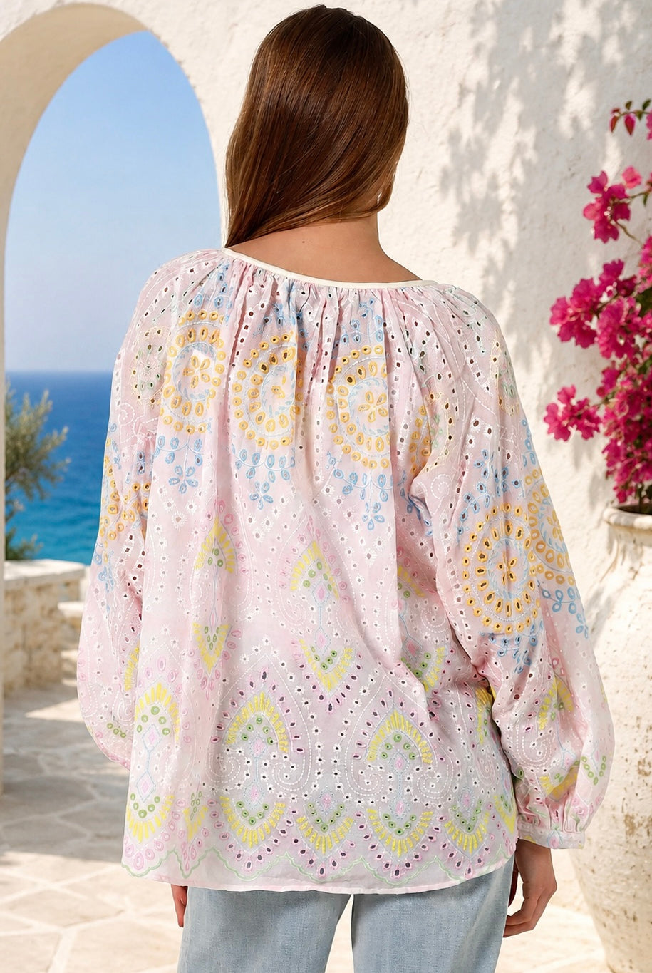 A woman viewed from behind wearing a light pink eyelet blouse with yellow, blue, and green bohemian circular patterns, set against a Mediterranean-style background with a blue sea and white stucco wall.