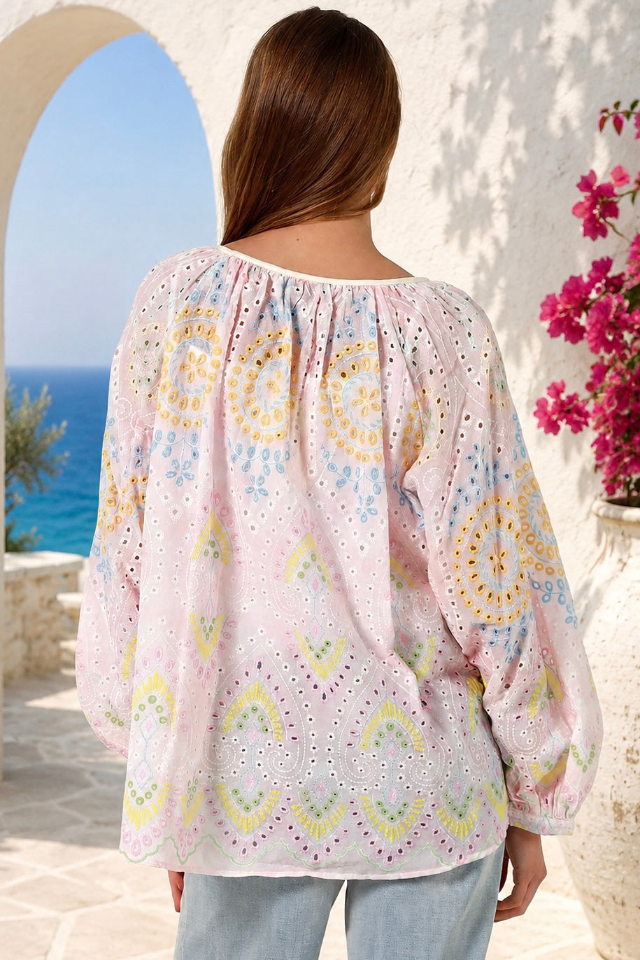 A woman viewed from behind wearing a light pink eyelet blouse with yellow, blue, and green bohemian circular patterns, set against a Mediterranean-style background with a blue sea and white stucco wall.
