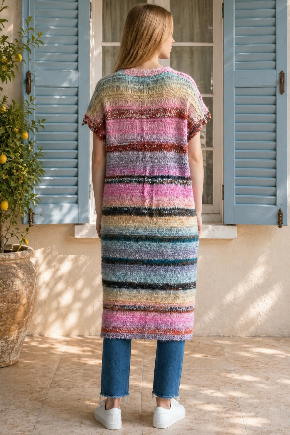 A woman wearing a long, open-front, short-sleeved chunky knit cardigan with horizontal multicolor stripes in pink, blue, beige, brown, and white, paired with a white tank top and blue jeans, standing in front of a window with blue shutters.