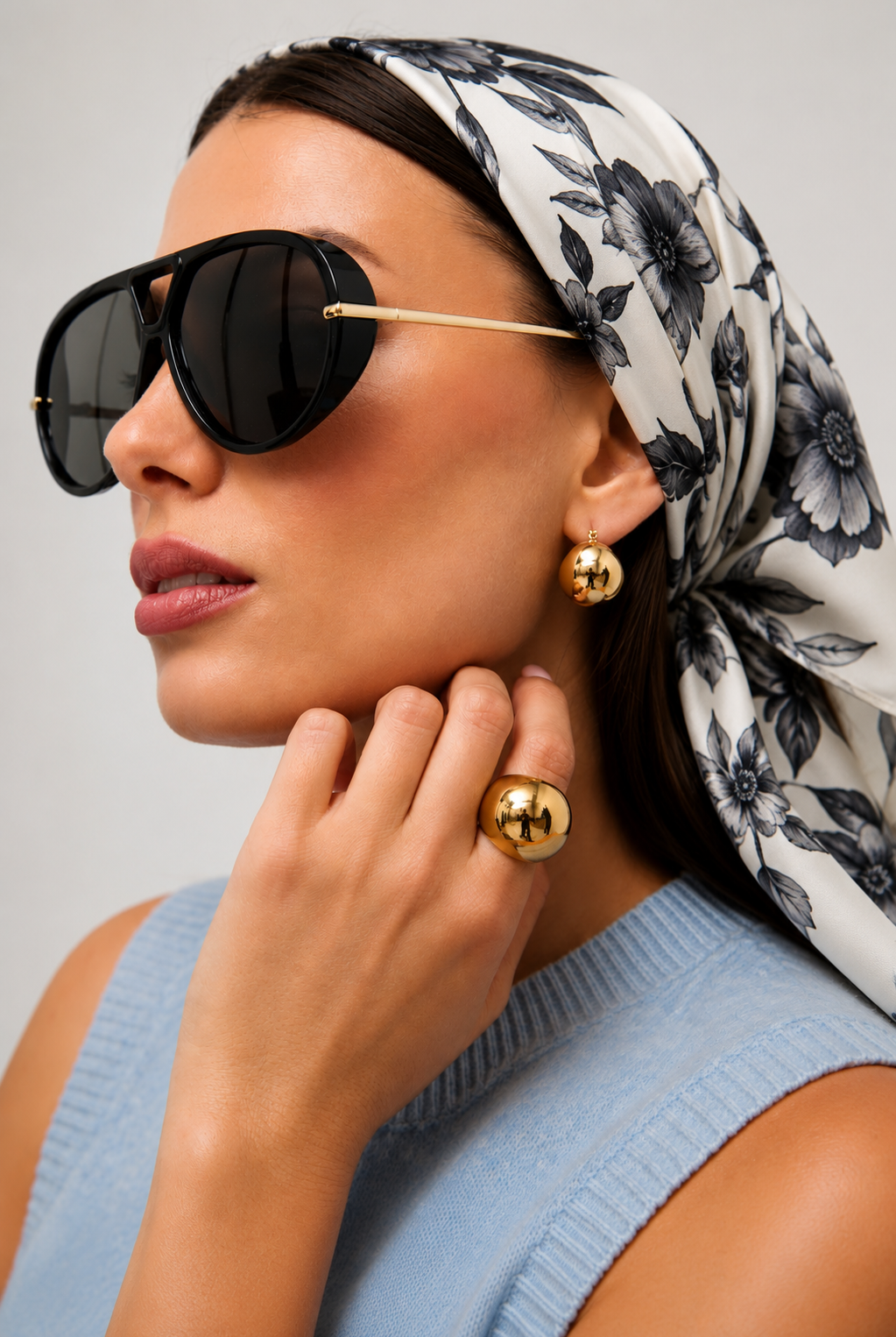 A close-up of a woman wearing oversized black aviator-style sunglasses with gold metal temples, along with gold hoop earrings and gold rings, against a light gray background.