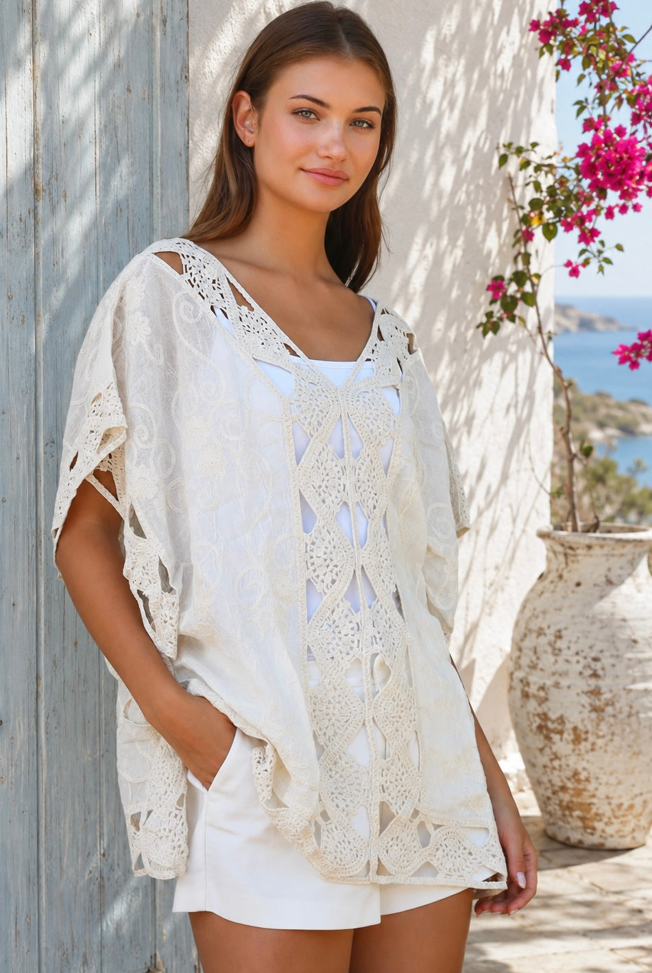 Cream crochet boho kaftan with ornate lace panels and relaxed sleeves in a sunlit Mediterranean coastal setting.