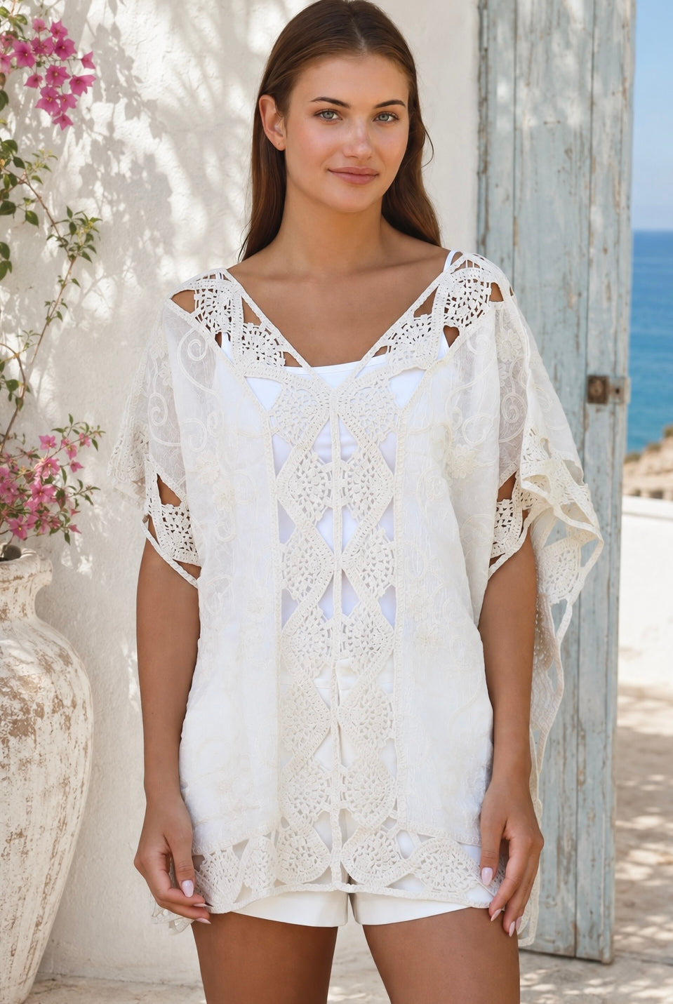 Cream crochet boho kaftan with ornate lace panels and relaxed sleeves in a sunlit Mediterranean coastal setting.