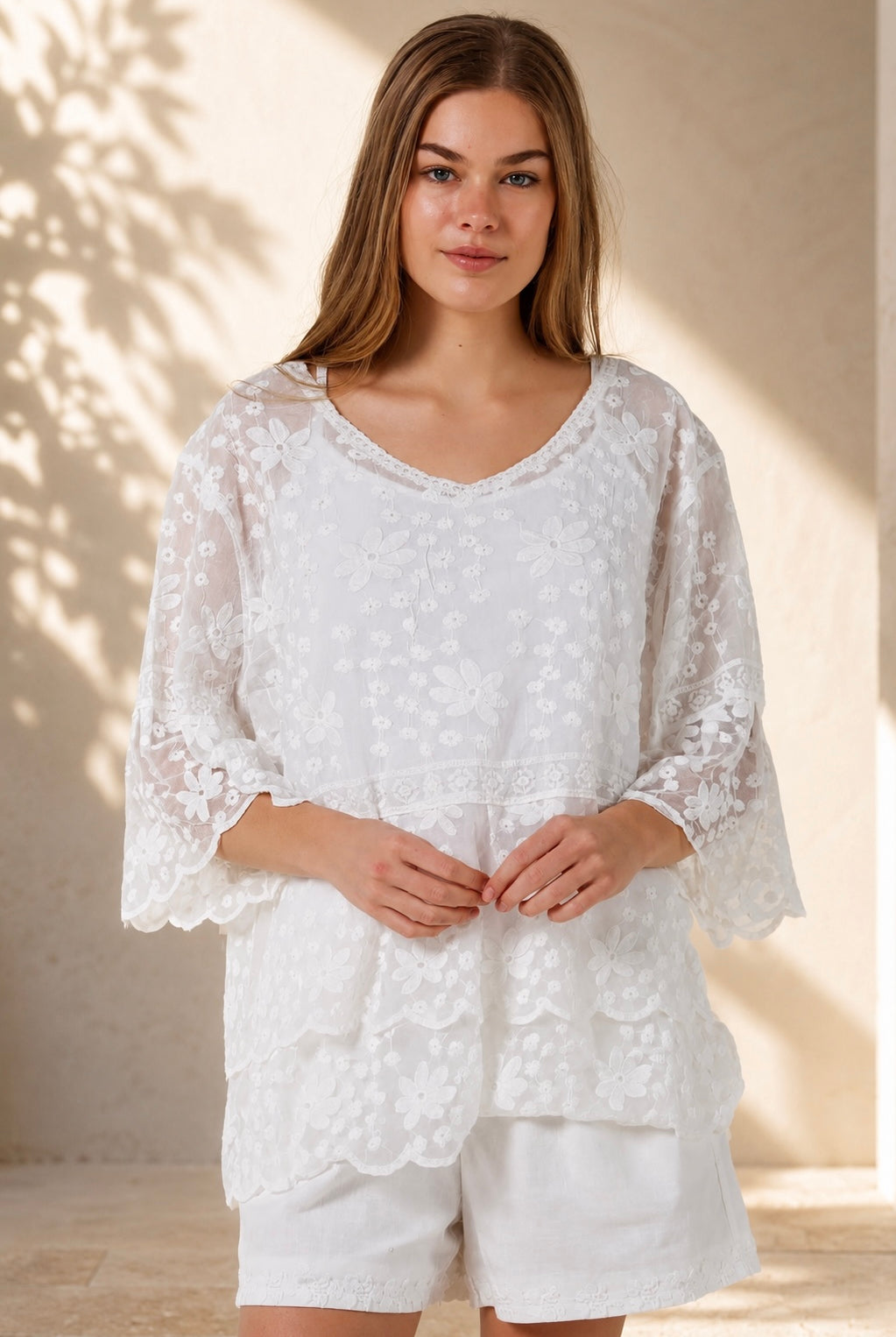 A woman wearing a white lace long-sleeve tunic top with matching white shorts, standing in a sunlit outdoor setting with a beige wall and potted plant in the background.