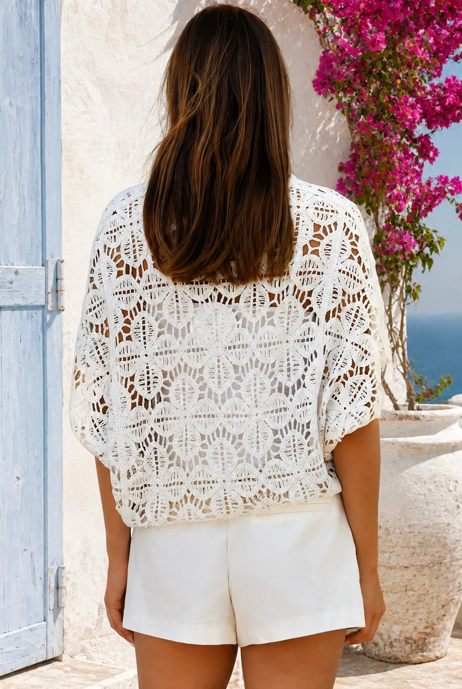 A woman viewed from behind wearing a white crochet lace top and white shorts, standing outdoors against a white wall with a blue door and pink bougainvillea flowers.