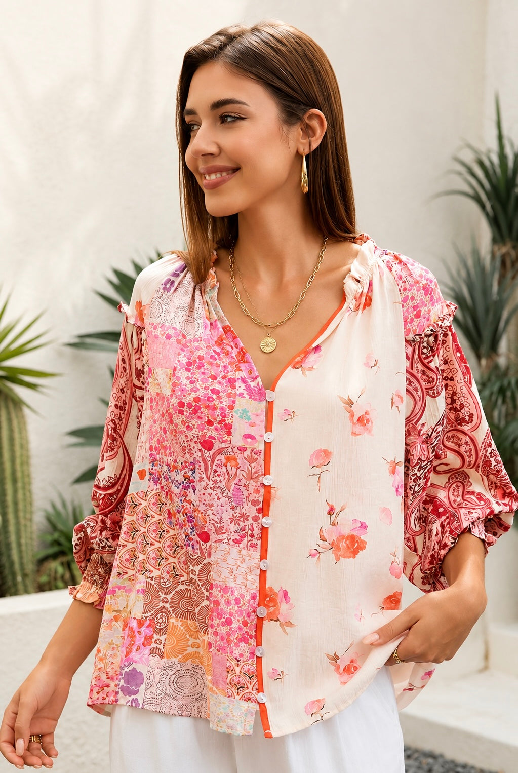 A woman wearing a bohemian-style long-sleeve blouse with a pink, red, and orange floral patchwork print on a white base, styled with white pants and gold jewelry in an outdoor setting.