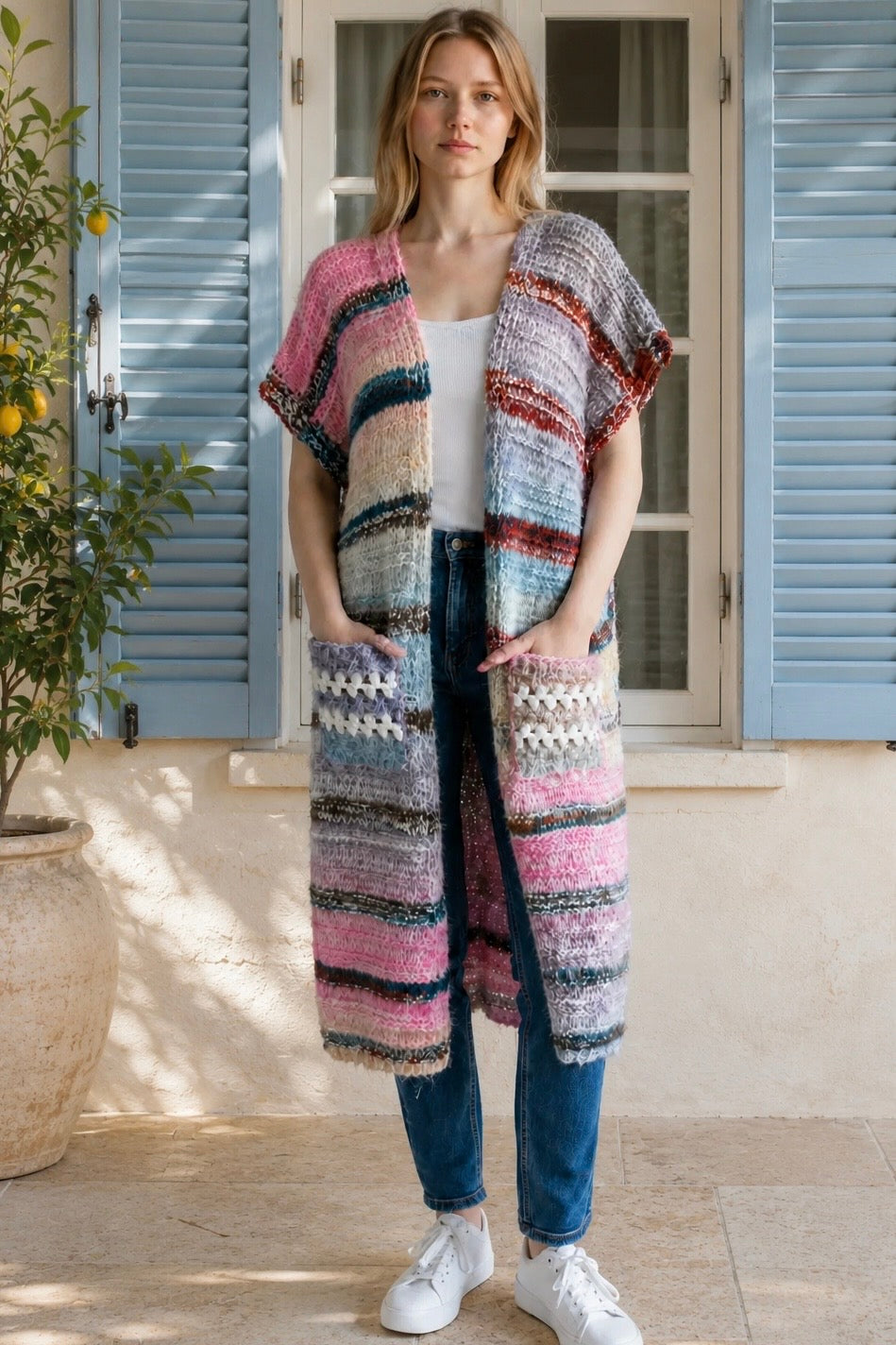 A woman wearing a long, open-front, short-sleeved chunky knit cardigan with horizontal multicolor stripes in pink, blue, beige, brown, and white, paired with a white tank top and blue jeans, standing in front of a window with blue shutters.