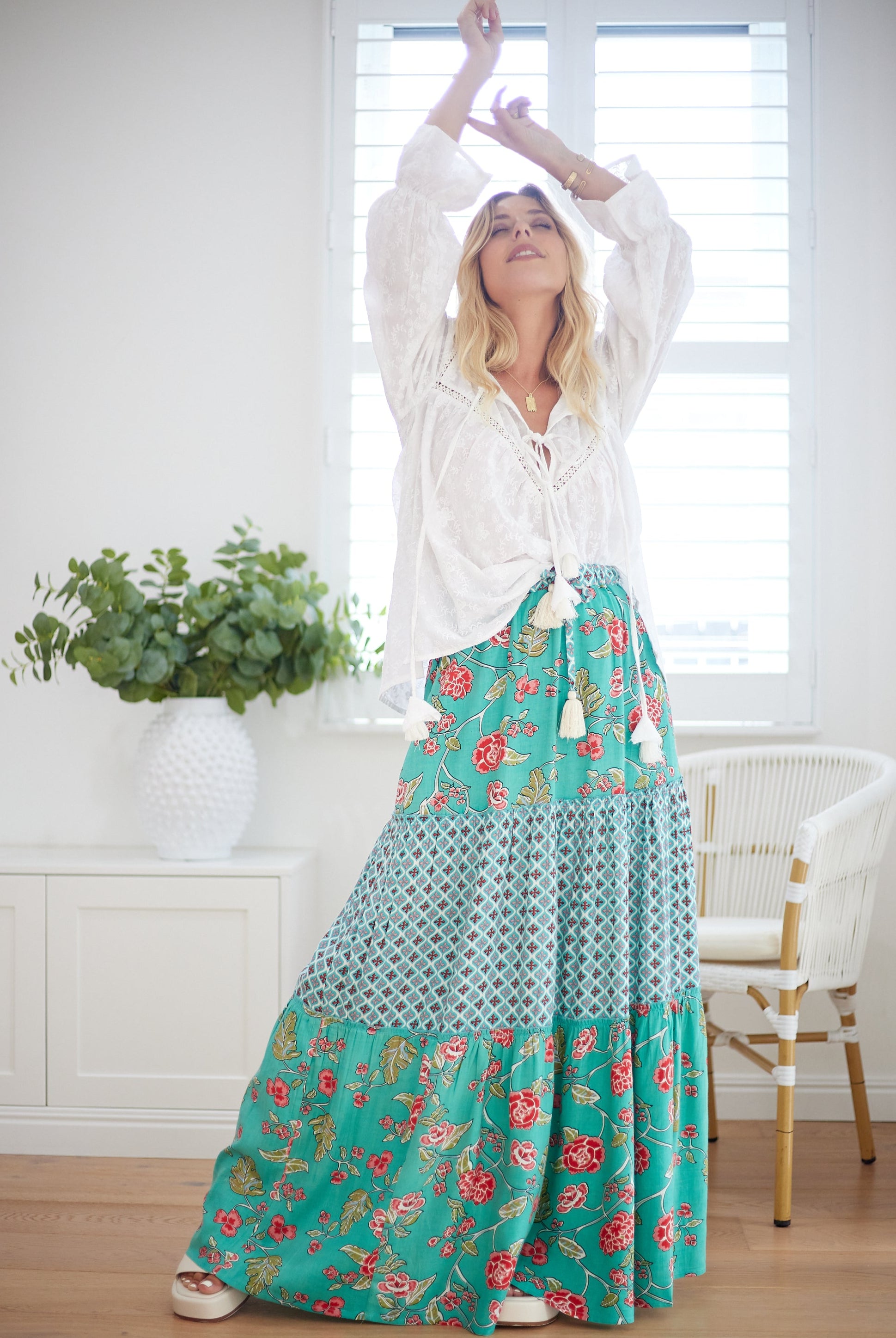 Woman wearing a white blouse and a long turquoise floral skirt in a bright room.