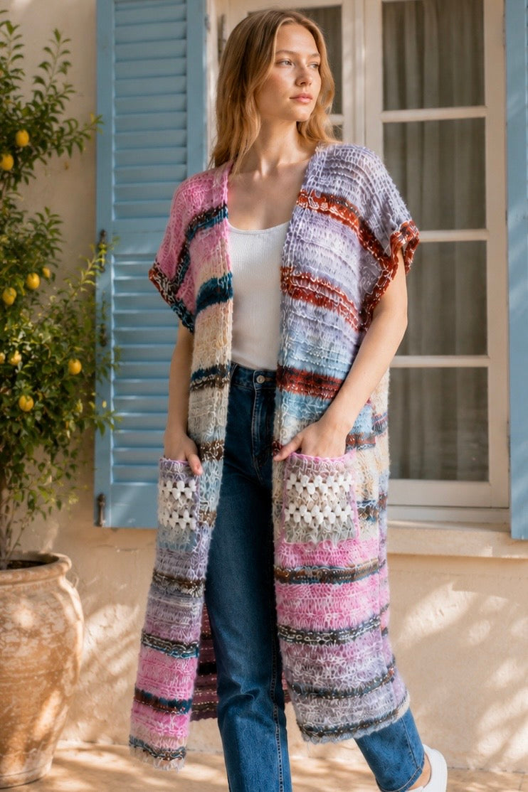 A woman wearing a long, open-front, short-sleeved chunky knit cardigan with horizontal multicolor stripes in pink, blue, beige, brown, and white, paired with a white tank top and blue jeans, standing in front of a window with blue shutters.