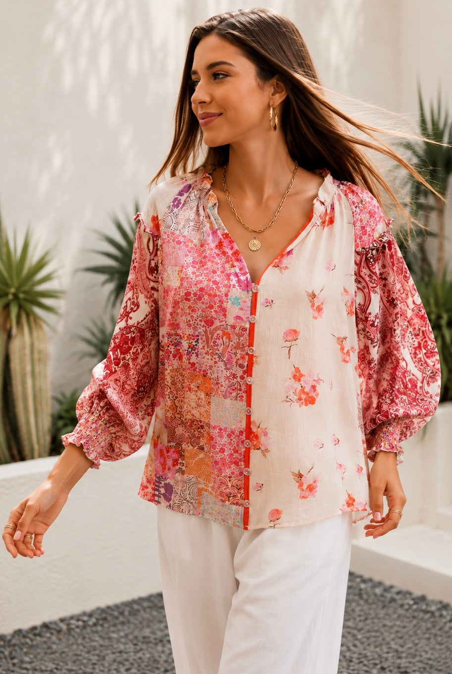 A woman wearing a bohemian-style long-sleeve blouse with a pink, red, and orange floral patchwork print on a white base, styled with white pants and gold jewelry in an outdoor setting.
