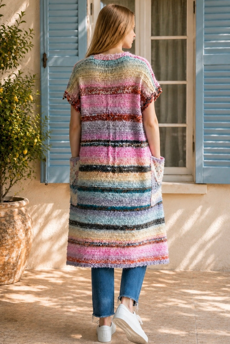 A woman wearing a long, open-front, short-sleeved chunky knit cardigan with horizontal multicolor stripes in pink, blue, beige, brown, and white, paired with a white tank top and blue jeans, standing in front of a window with blue shutters.