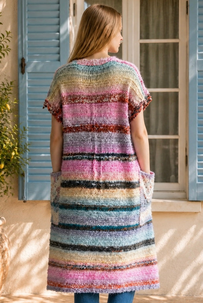 A woman wearing a long, open-front, short-sleeved chunky knit cardigan with horizontal multicolor stripes in pink, blue, beige, brown, and white, paired with a white tank top and blue jeans, standing in front of a window with blue shutters.