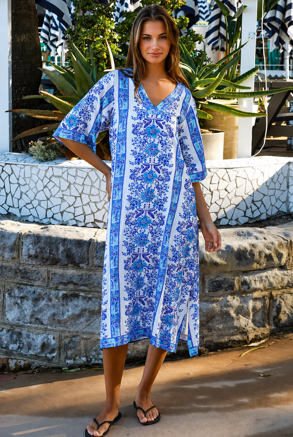 A woman wearing a blue and white patterned kaftan dress with a V-neck and three-quarter sleeves, walking outdoors in front of a stone wall and tropical plants.