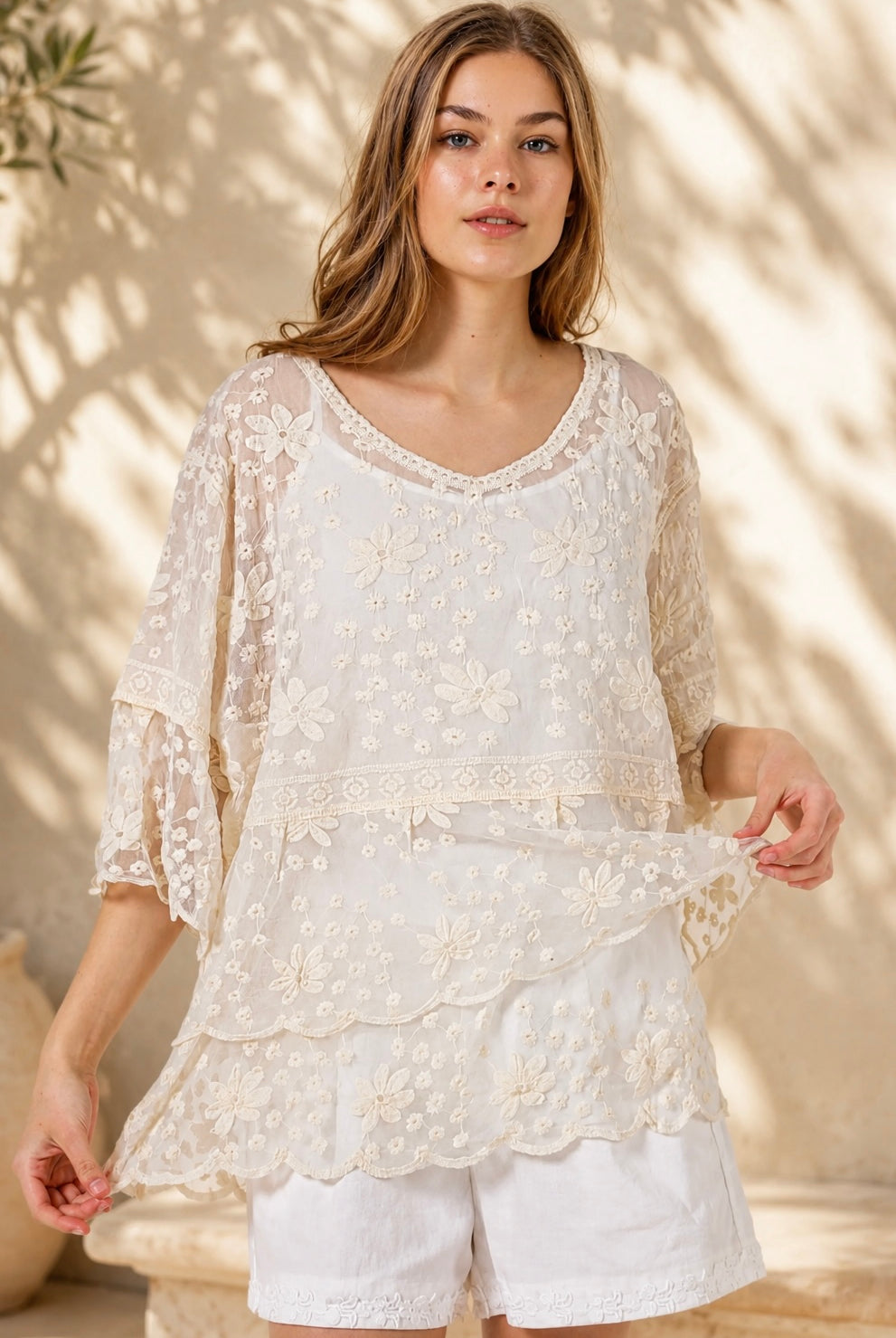 A woman wearing a cream lace top with three-quarter sleeves and white shorts, standing in front of a warm beige wall with leaf shadows.