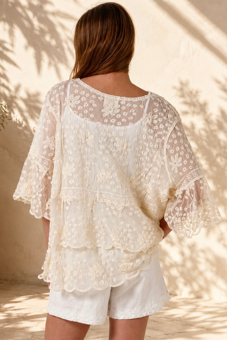 A woman wearing a cream lace top with three-quarter sleeves and white shorts, standing in front of a warm beige wall with leaf shadows.