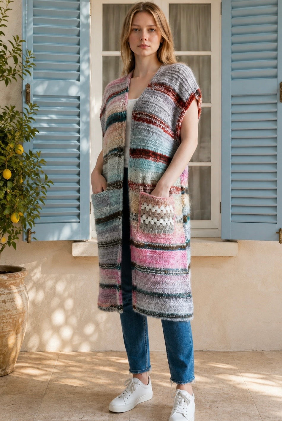 A woman wearing a long, open-front, short-sleeved chunky knit cardigan with horizontal multicolor stripes in pink, blue, beige, brown, and white, paired with a white tank top and blue jeans, standing in front of a window with blue shutters.