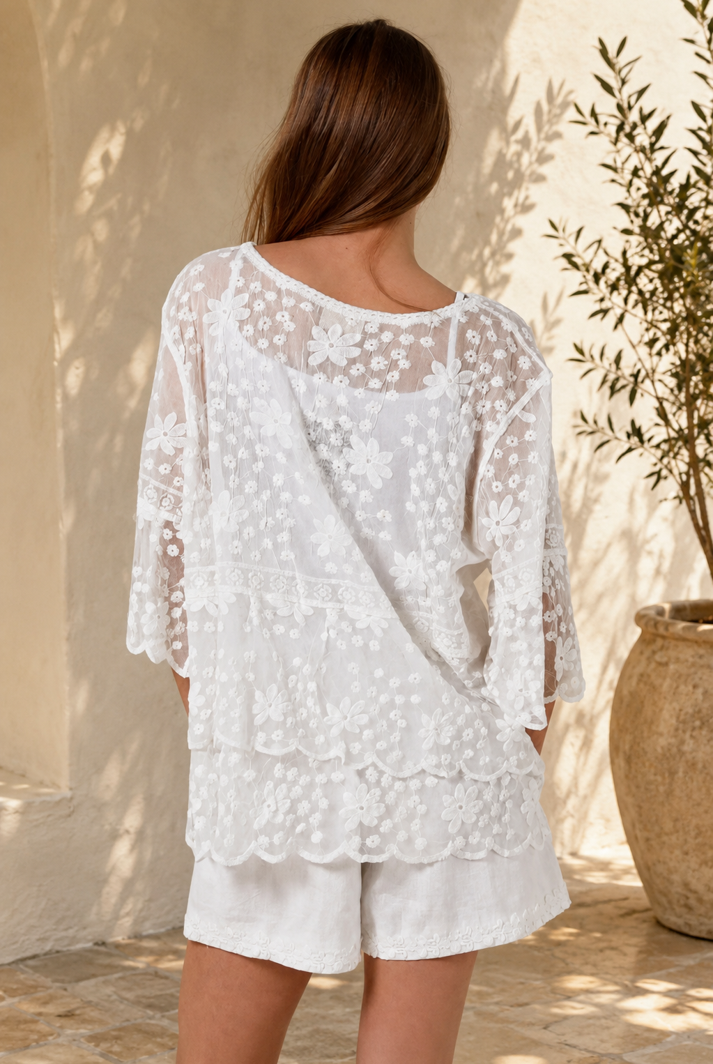 A woman wearing a white lace long-sleeve tunic top with matching white shorts, standing in a sunlit outdoor setting with a beige wall and potted plant in the background.