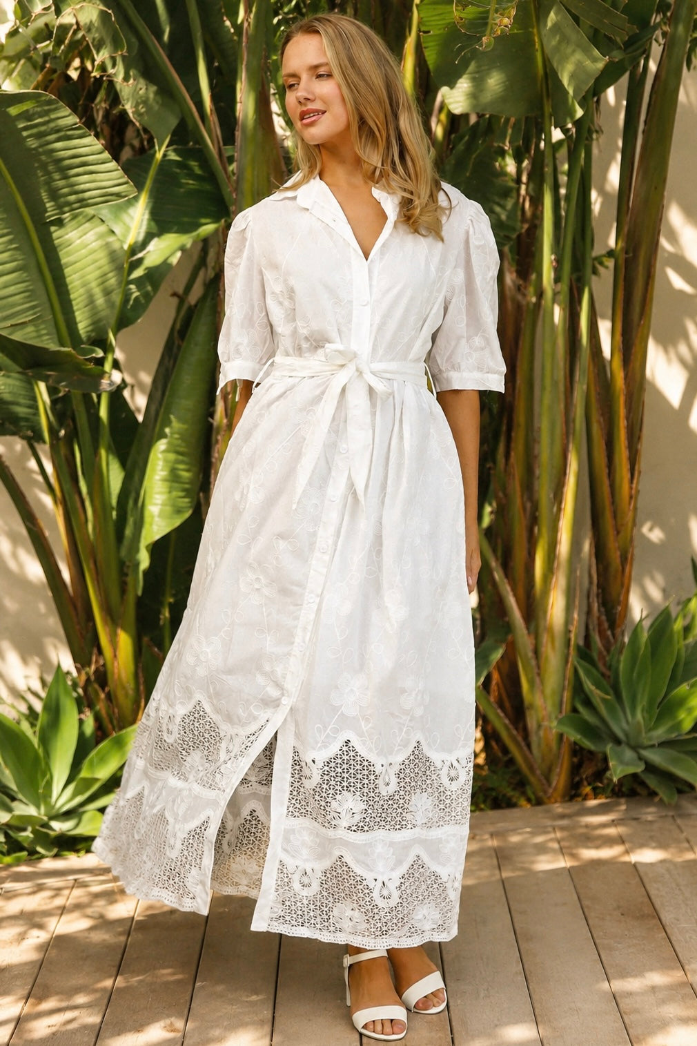 A woman wearing a white lace midi shirt dress with short puff sleeves, a tie waist, and a front slit, standing outdoors in front of tropical green foliage.