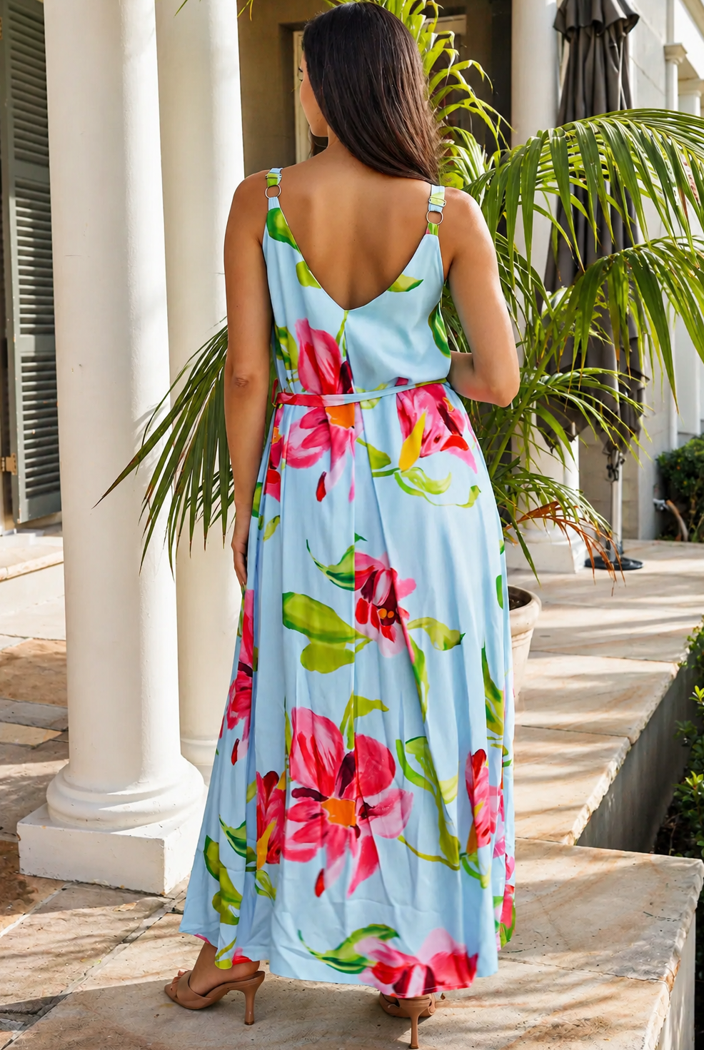 A woman viewed from behind wearing a light blue sleeveless maxi dress with a vibrant pink and green floral print, standing on a stone patio in front of white columns and palm trees.