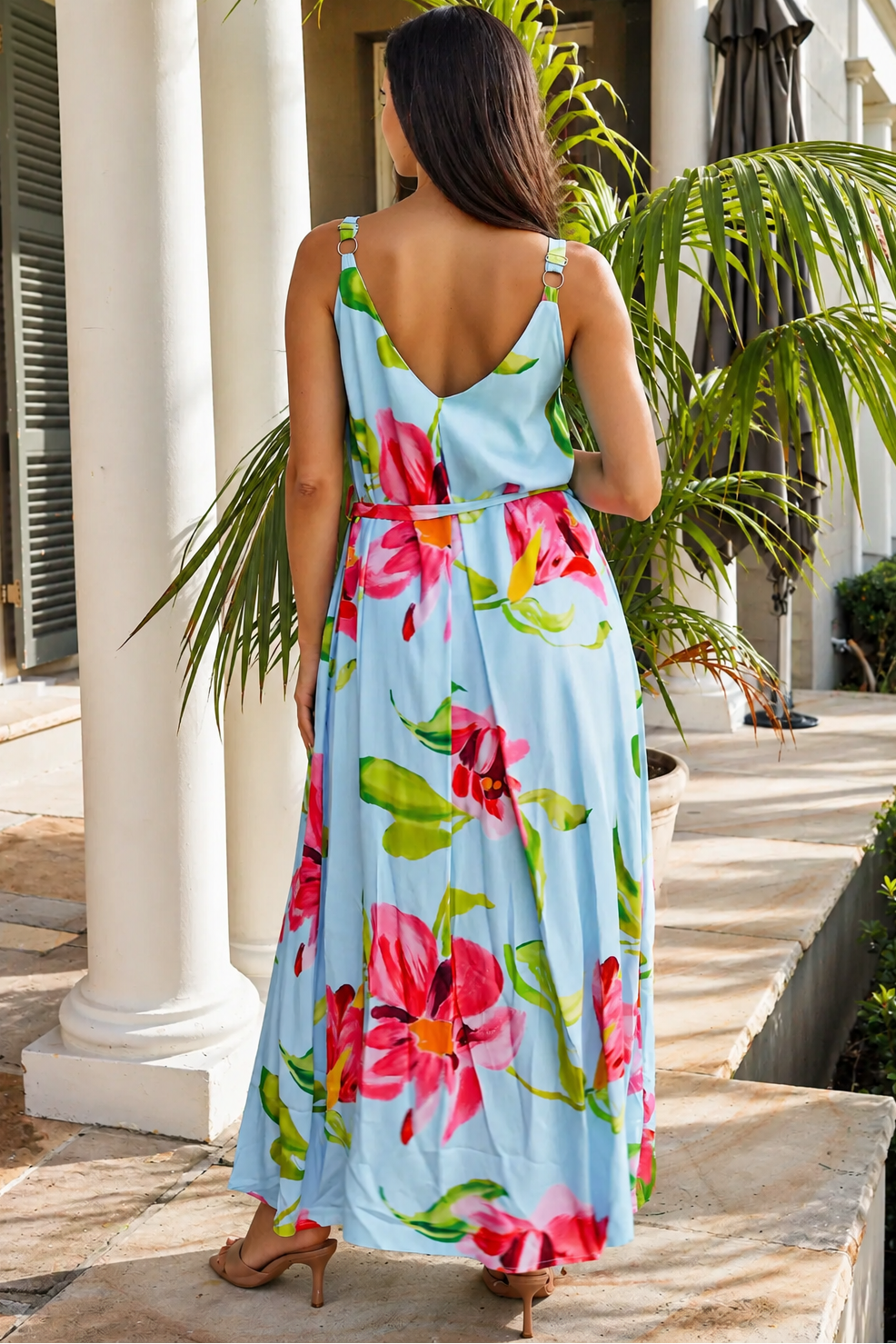 A woman viewed from behind wearing a light blue sleeveless maxi dress with a vibrant pink and green floral print, standing on a stone patio in front of white columns and palm trees.