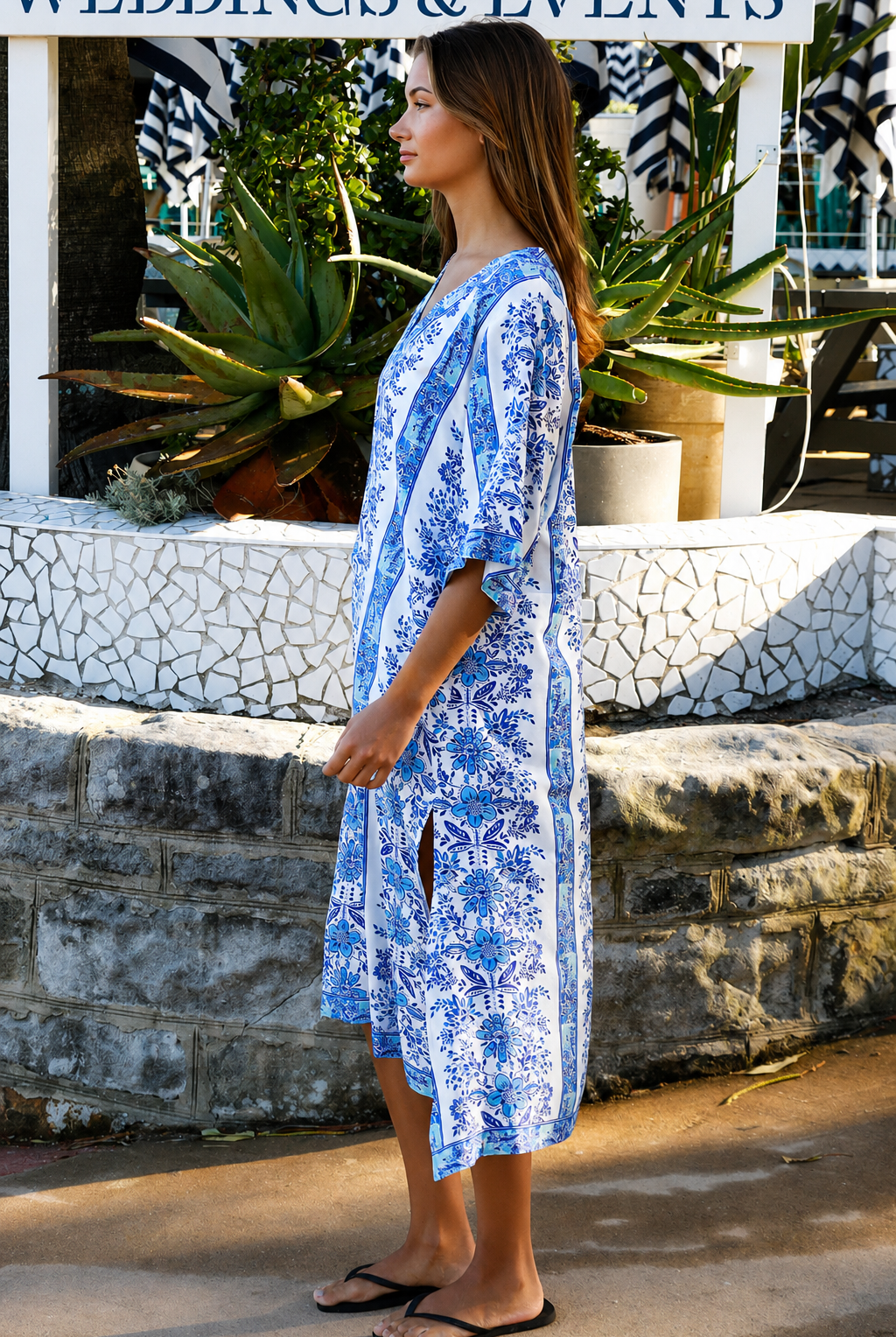 A woman wearing a blue and white patterned kaftan dress with a V-neck and three-quarter sleeves, walking outdoors in front of a stone wall and tropical plants.