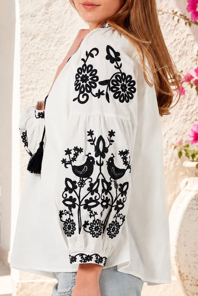 A woman wearing a white long-sleeve blouse with black floral and bird embroidery, paired with light blue jeans, photographed against a textured stucco wall with flowers in the background. Bohemian Love Runway.