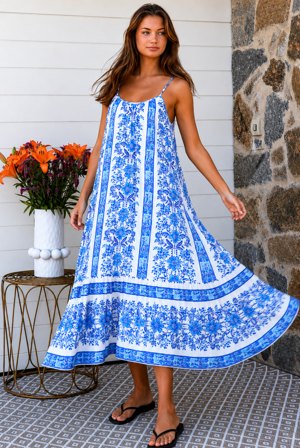 A woman modeling a blue and white patterned maxi dress with spaghetti straps, standing on a patterned tile porch next to a small table with orange flowers.