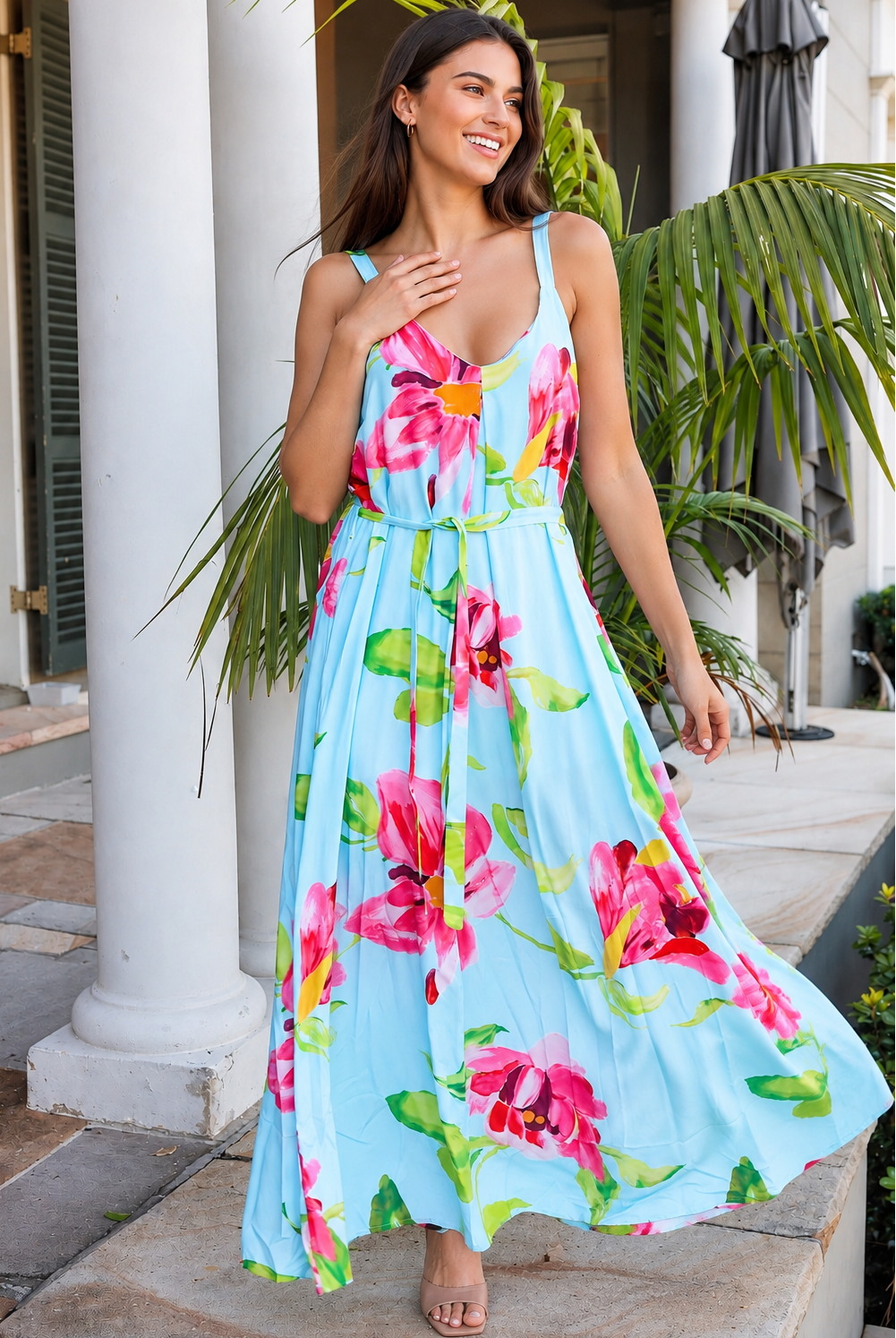 A woman wearing a light blue sleeveless maxi dress with a vibrant pink and green floral print, standing outdoors in front of white columns and tropical plants.