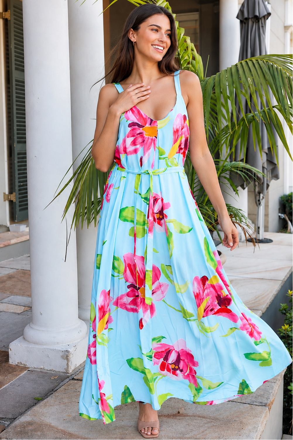 A woman wearing a light blue sleeveless maxi dress with a vibrant pink and green floral print, standing outdoors in front of white columns and tropical plants.
