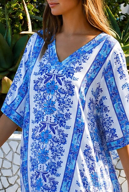 A woman wearing a white kaftan-style tunic dress with a vibrant blue floral and geometric pattern, photographed outdoors with green foliage in the background.