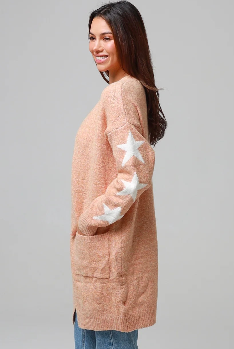 Woman wearing a peachy-blush longline cardigan with white star accents, front pockets, and relaxed fit — cozy knitwear styled with jeans and a white top, perfect for layering.
