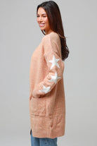 Woman wearing a peachy-blush longline cardigan with white star accents, front pockets, and relaxed fit — cozy knitwear styled with jeans and a white top, perfect for layering.
