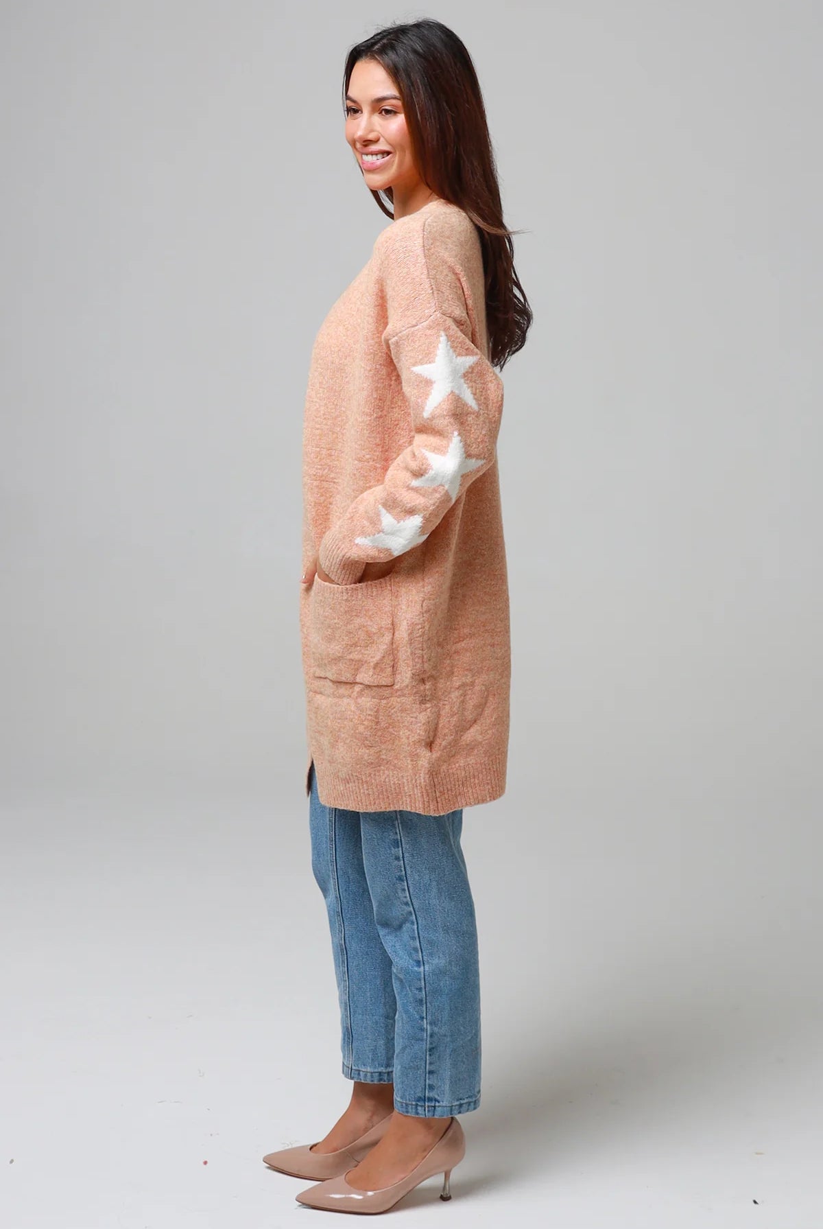 Woman wearing a peachy-blush longline cardigan with white star accents, front pockets, and relaxed fit — cozy knitwear styled with jeans and a white top, perfect for layering.