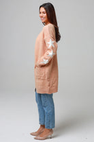 Woman wearing a peachy-blush longline cardigan with white star accents, front pockets, and relaxed fit — cozy knitwear styled with jeans and a white top, perfect for layering.