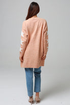 Woman wearing a peachy-blush longline cardigan with white star accents, front pockets, and relaxed fit — cozy knitwear styled with jeans and a white top, perfect for layering.