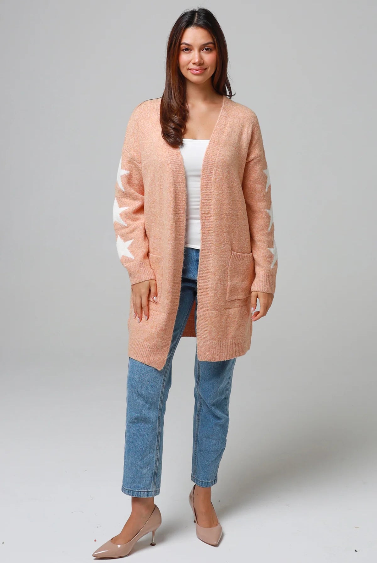 Woman wearing a peachy-blush longline cardigan with white star accents, front pockets, and relaxed fit — cozy knitwear styled with jeans and a white top, perfect for layering.
