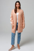 Woman wearing a peachy-blush longline cardigan with white star accents, front pockets, and relaxed fit — cozy knitwear styled with jeans and a white top, perfect for layering.