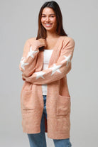Woman wearing a peachy-blush longline cardigan with white star accents, front pockets, and relaxed fit — cozy knitwear styled with jeans and a white top, perfect for layering.