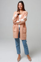Woman wearing a peachy-blush longline cardigan with white star accents, front pockets, and relaxed fit — cozy knitwear styled with jeans and a white top, perfect for layering.