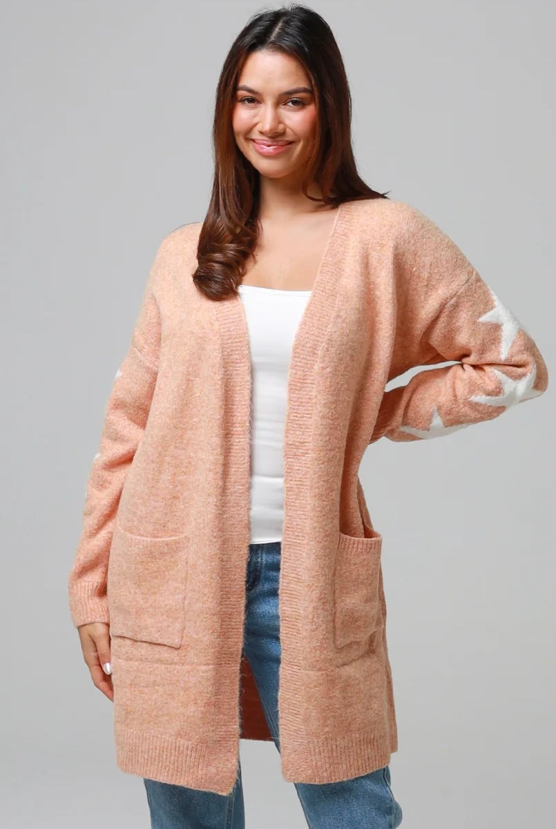 Woman wearing a peachy-blush longline cardigan with white star accents, front pockets, and relaxed fit — cozy knitwear styled with jeans and a white top, perfect for layering.