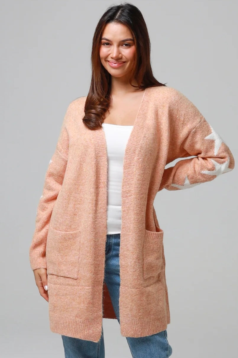 Woman wearing a peachy-blush longline cardigan with white star accents, front pockets, and relaxed fit — cozy knitwear styled with jeans and a white top, perfect for layering.