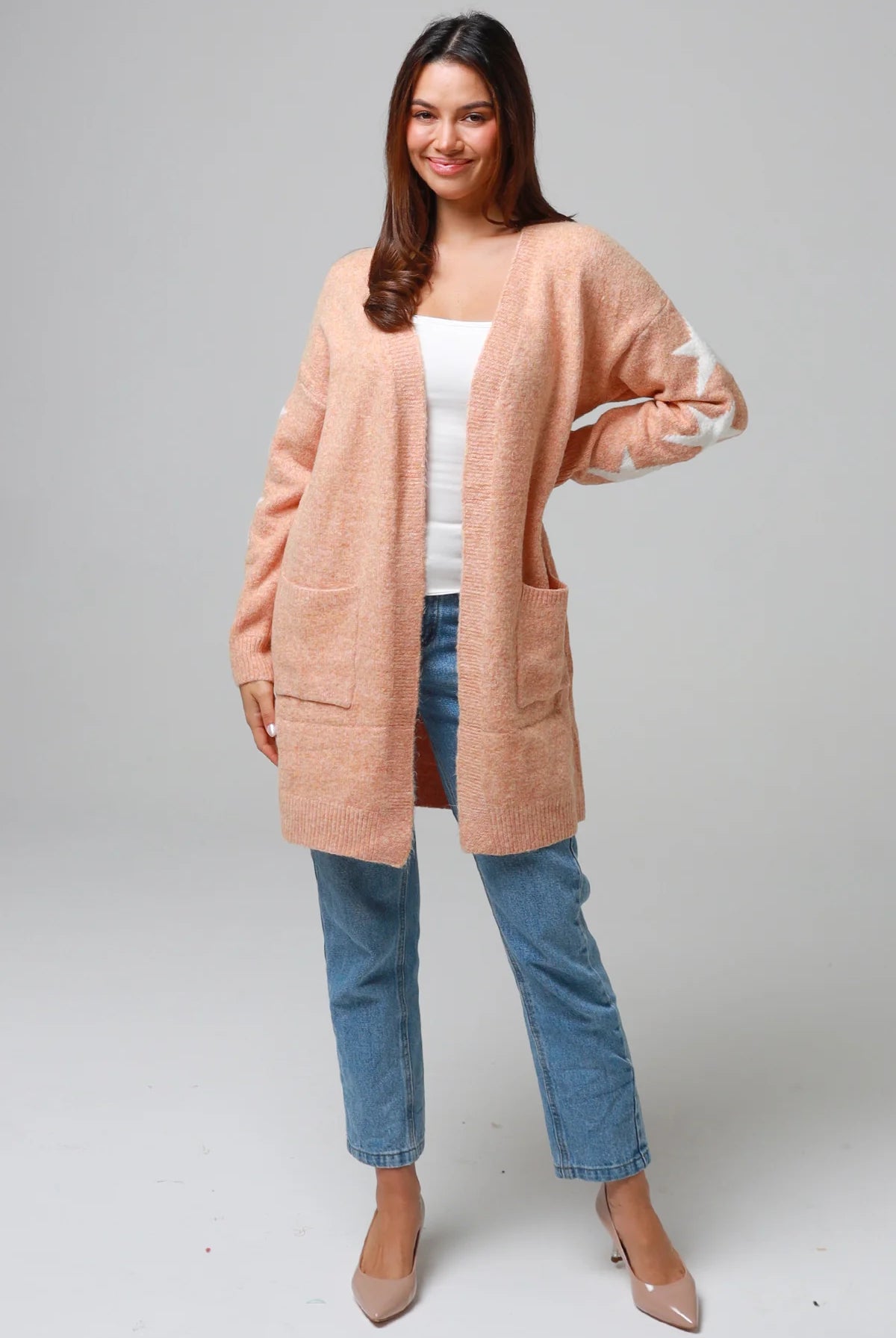 Woman wearing a peachy-blush longline cardigan with white star accents, front pockets, and relaxed fit — cozy knitwear styled with jeans and a white top, perfect for layering.